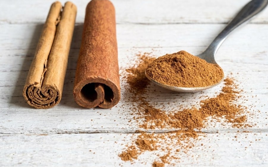 Three cinnamon quills on white painted boards beside a spoonful of ground cinnamon, with powder scattered across the wood